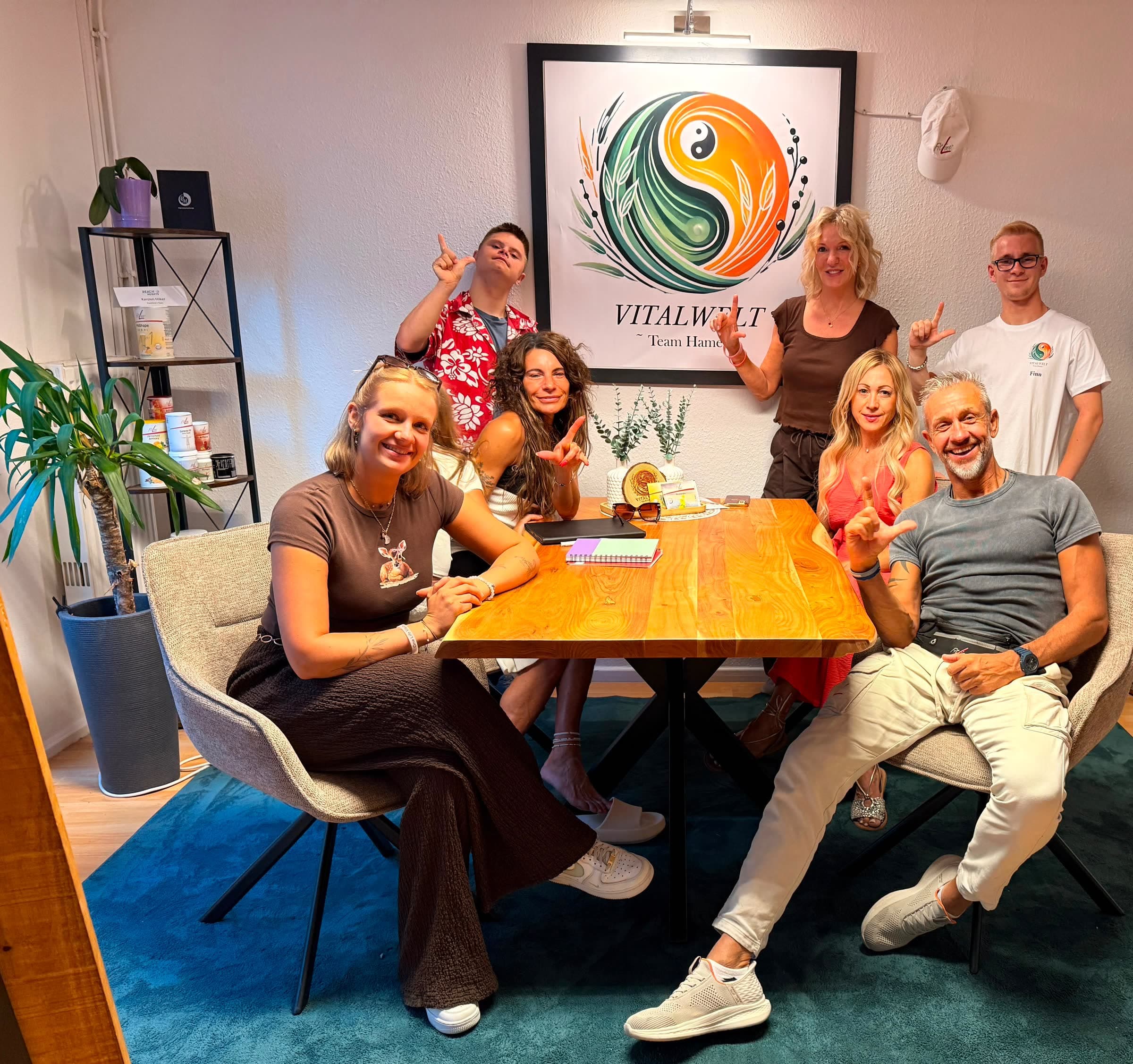 Das VitalWelt Team Hameln in den eigenen Räumen in der Baustraße 8, 31785 Hameln: das Team sitzt und steht um den Besprechungstisch unter dem gerahmten VitalWelt-Logo.